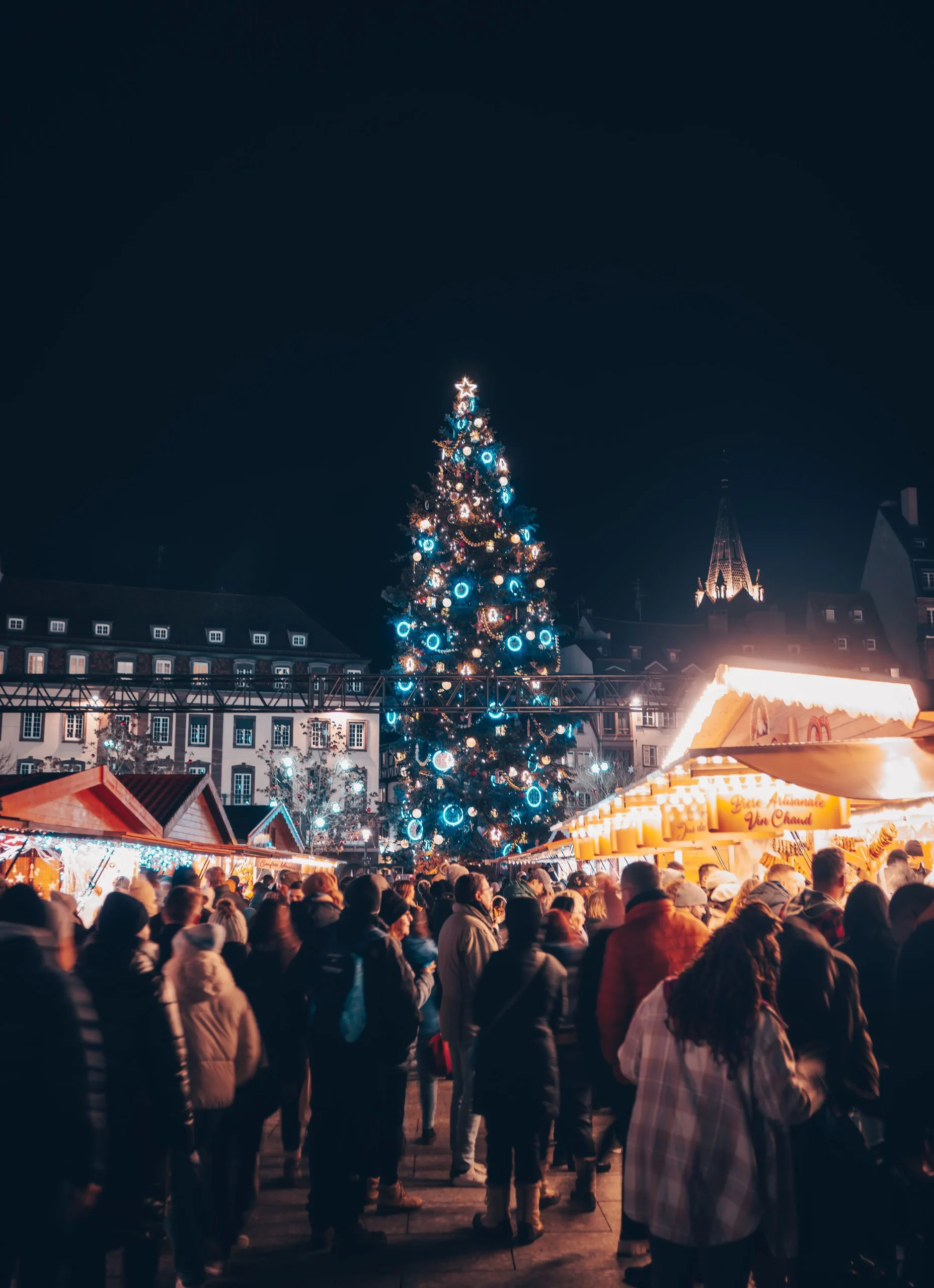 Une foule rassemblée au marché de Noël de Strasbourg de nuit, dominé par un grand sapin illuminé de lumières bleues et blanches, entouré de chalets en bois festifs et de visiteurs emmitouflés.