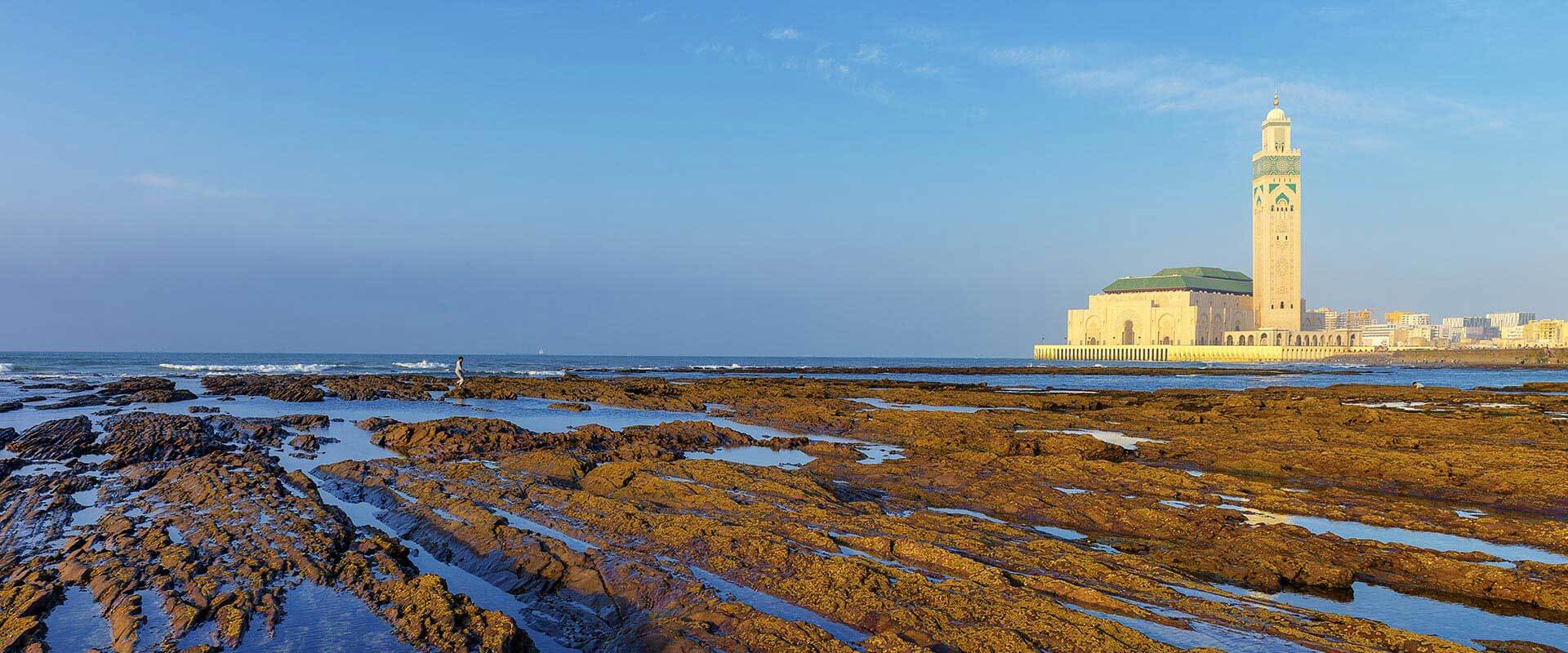 Vue de la Mosquée Hassan II de Casablanca depuis la rive, montrant des rochers à marée basse et l'océan Atlantique en arrière-plan.