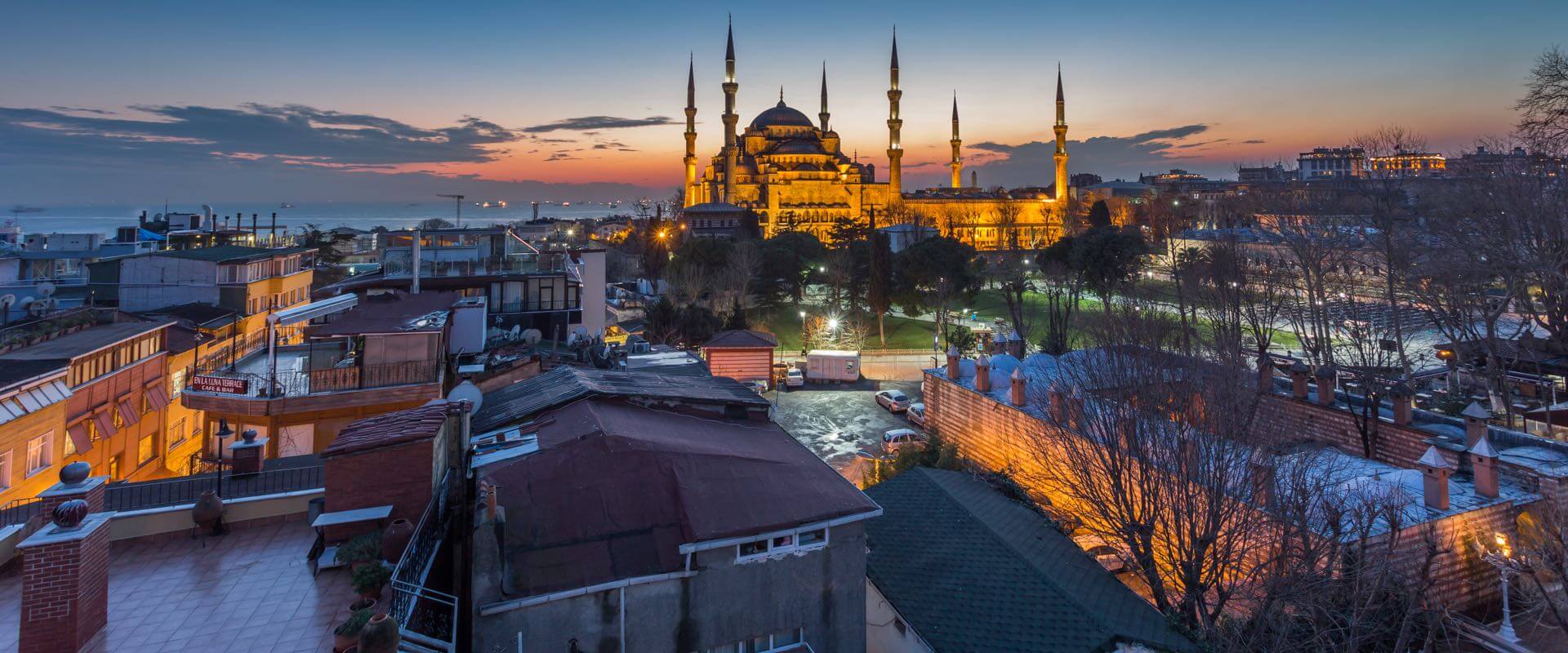 Vue panoramique de la Mosquée Bleue illuminée au crépuscule à Istanbul, depuis une zone résidentielle avec des toits et des arbres visibles.