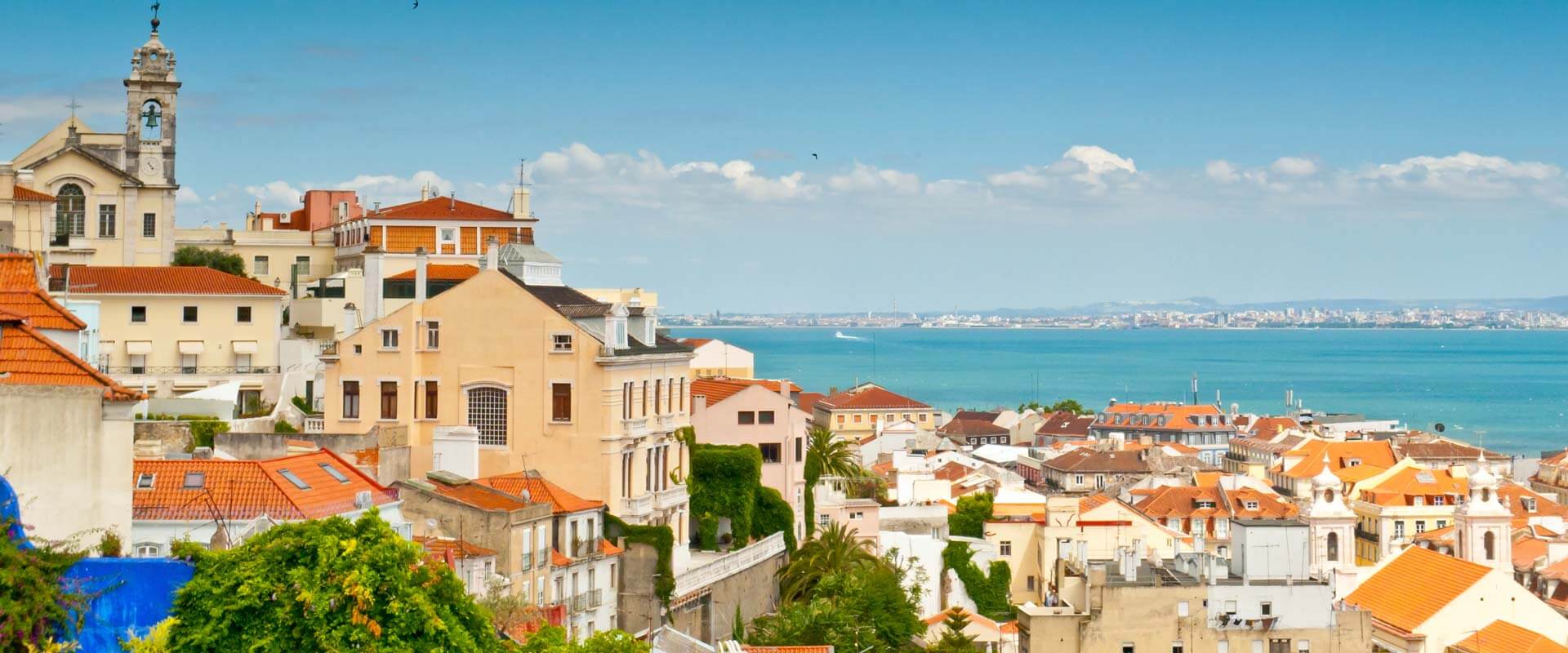 Vue panoramique de Lisbonne avec des maisons colorées et des toits rouges, une église avec un clocher, des arbres verts et le Tage en arrière-plan.