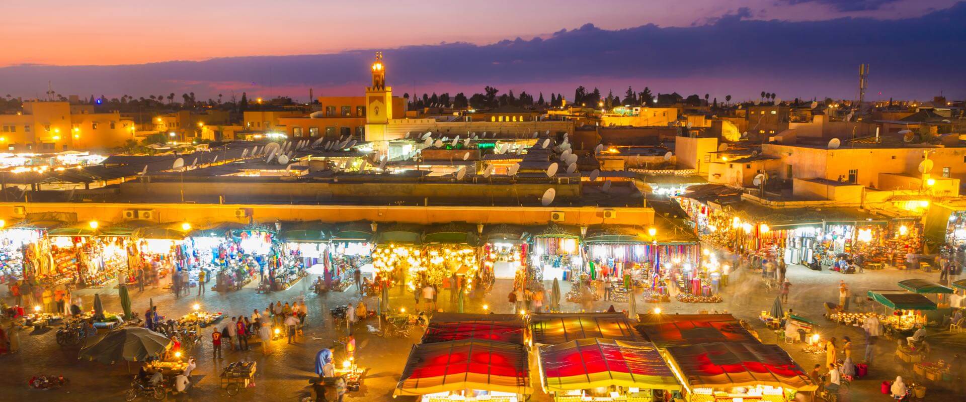 Vue aérienne nocturne de la place Jamaa el-Fna à Marrakech, Maroc, avec des stands de nourriture illuminés, des personnes qui se promènent et une mosquée avec un minaret en arrière-plan.